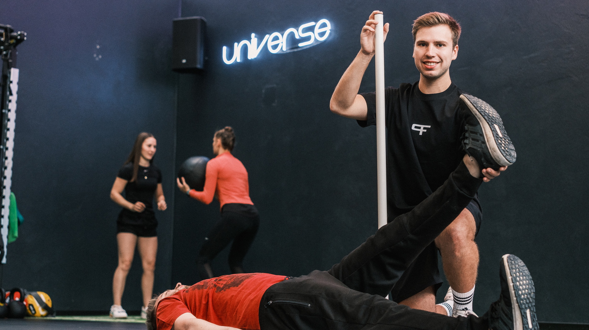 Ein Trainer führt eine Beweglichkeitsanalyse eines liegenden Mannes durch, im Hintergrund trainieren zwei Frauen mit einem Medizinball
