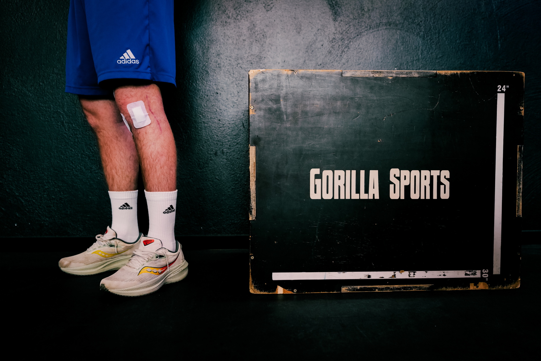 Eine Person steht vor einer Trainingsbox und trägt am Knie zwei frische Verbände nach einem operativen Eingriff.