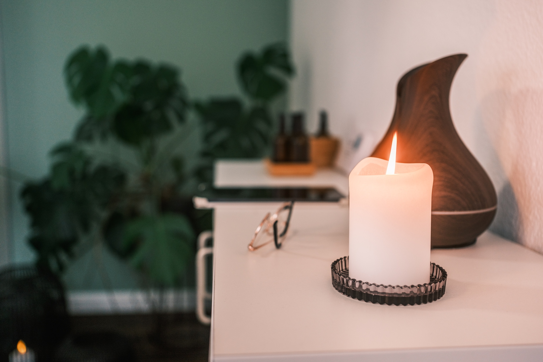ChatGPT:  Eine brennende Kerze steht auf einem Tisch und sorgt gemeinsam mit Pflanzen und einem Diffuser im Hintergrund für eine ruhige Wellnessatmosphäre.