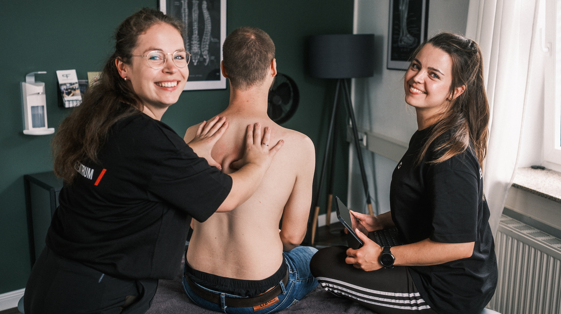 Zwei Therapeutinnen behandeln einen Patienten am Rücken, eine dokumentiert die Behandlung auf einem Tablet