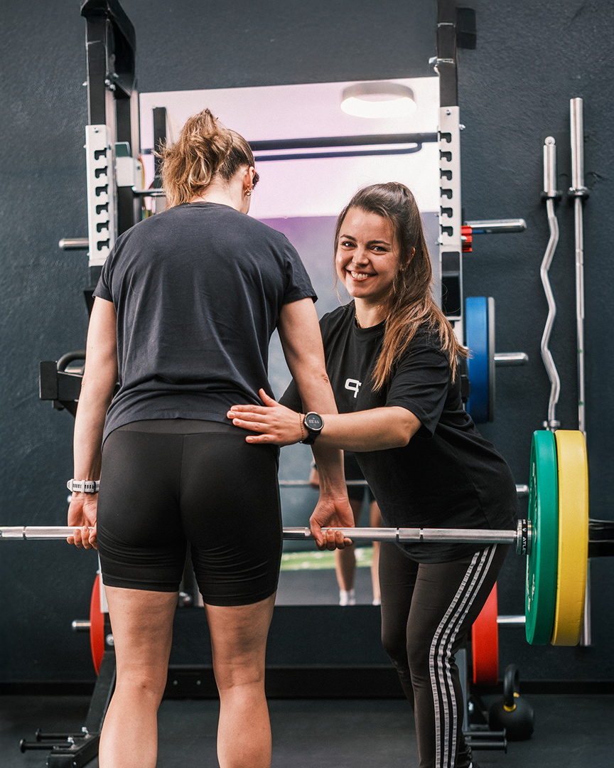 Eine Therapeutin unterstützt eine Patientin beim Kreuzheben mit der Langhantel.